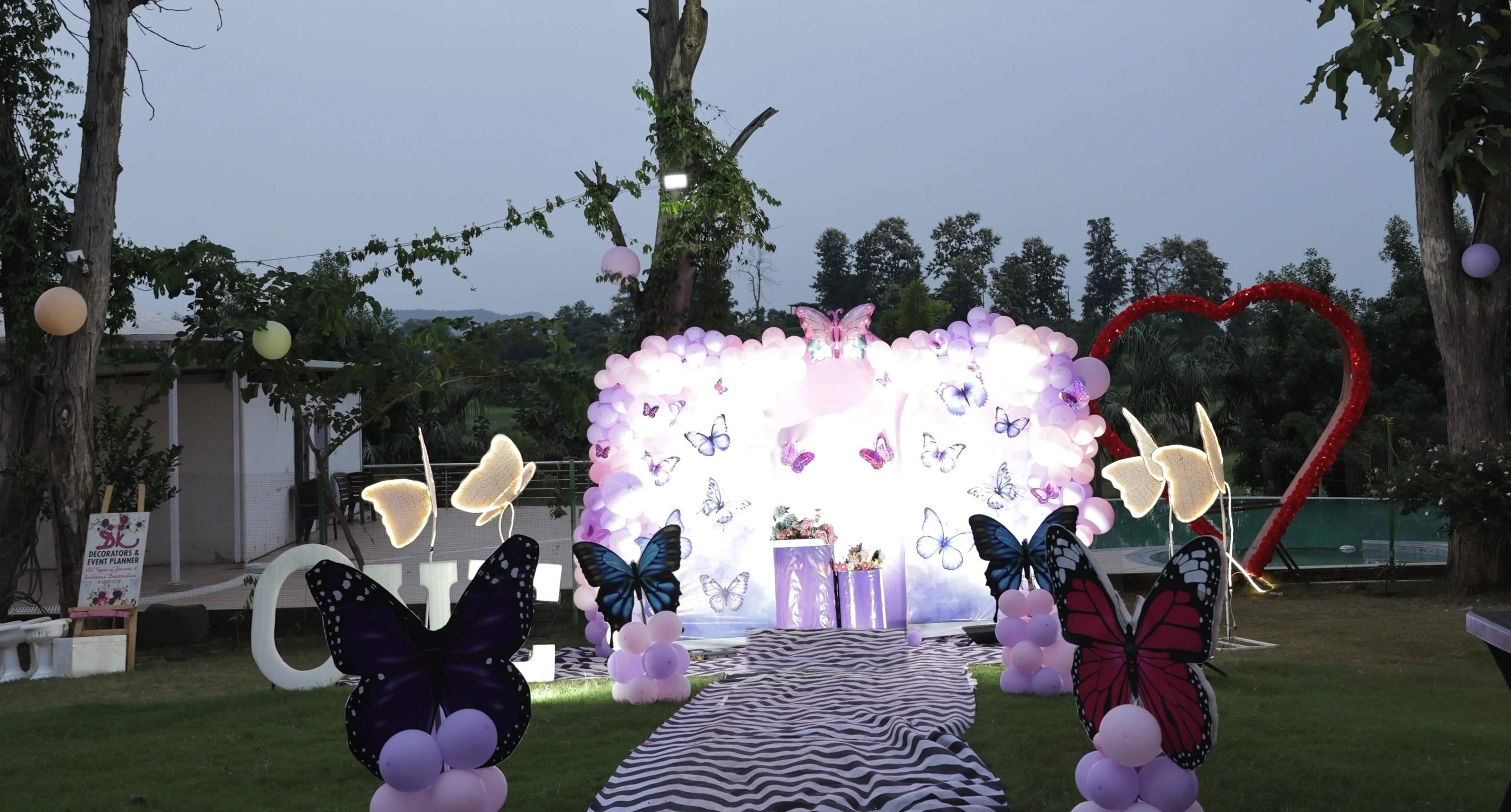 Birthday party on the lawn at dusk: a butterfly balloon stage, lit butterflies and a striped walkway under the trees