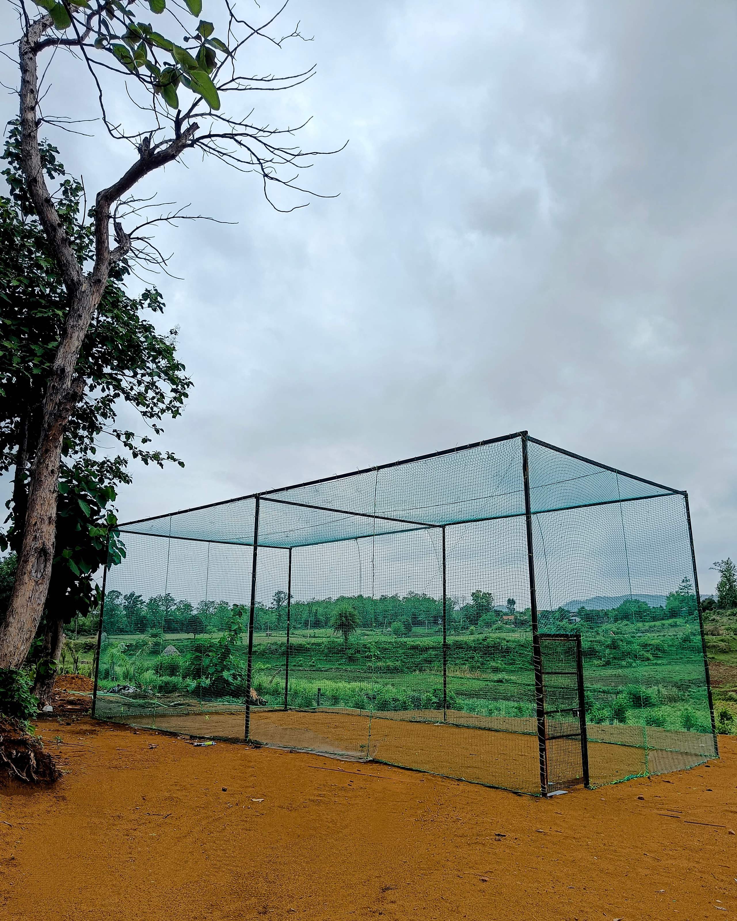 Cricket practice net on a red-earth pitch, with green fields behind