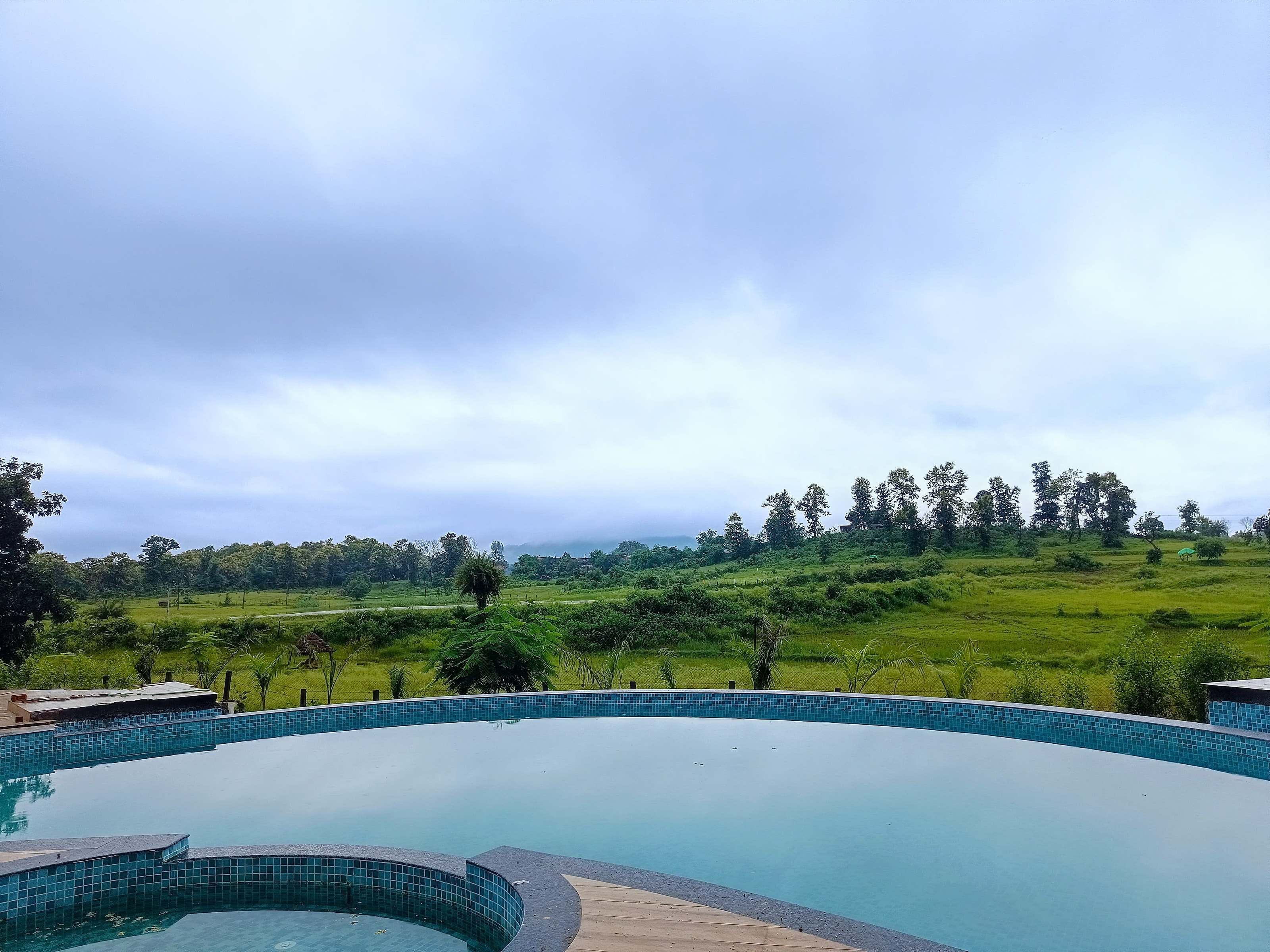 View across the pool edge to open green fields and distant hills under monsoon cloud